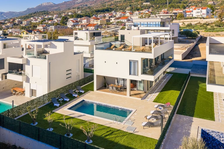Aerial view of modern white villa with swimming pool, manicured lawn, and mountain backdrop in Mediterranean hillside neighborhood