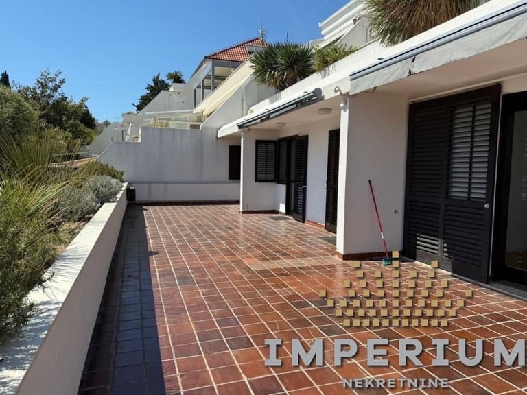 Modern Mediterranean-style home with terra cotta terrace, white walls, black shutters, and palm trees visible on a sunny day