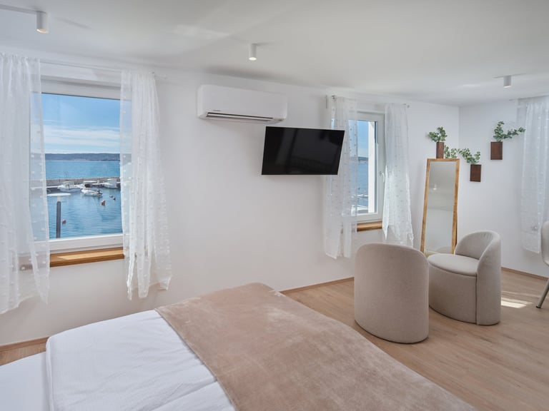 Bright modern bedroom with sea view, minimalist furnishings, white walls, wooden flooring, and a wall-mounted TV overlooking a coastal harbor