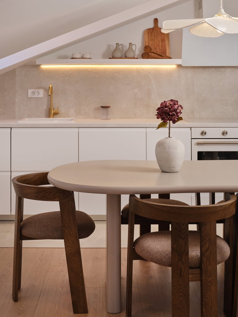 Modern kitchen dining area with round beige table, wooden chairs, white cabinetry, and stone backsplash with vase of flowers