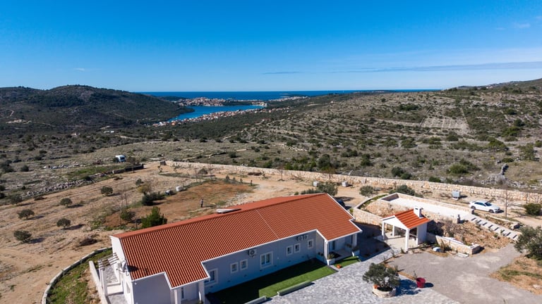 Aerial view of a blue and orange-roofed building on Mediterranean hillside overlooking coastal bay