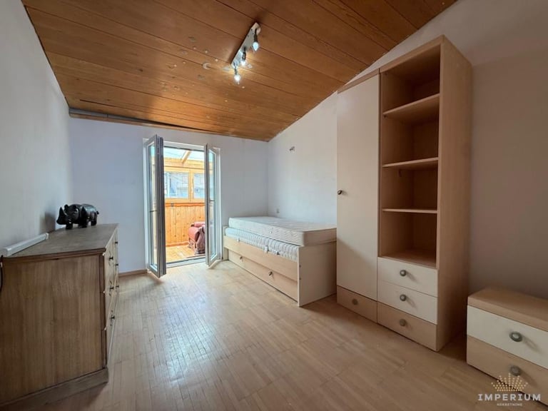 Bright bedroom with wooden ceiling, light wood furniture including shelving unit and dresser, open balcony door, and hardwood flooring.