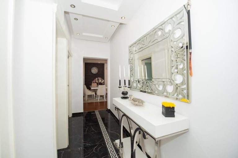 Modern white bathroom with ornate mirror, white vanity, black marble flooring, and doorway leading to bedroom