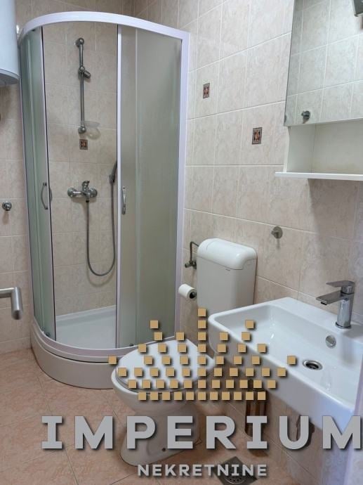 Modern bathroom with curved glass shower enclosure, sink, toilet, and small golden cube decorations arranged on floor with IMPERIUM NEKRETNINE text overlay