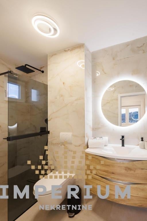 Modern bathroom with marble walls, circular illuminated mirror, floating wooden vanity, and glass shower enclosure