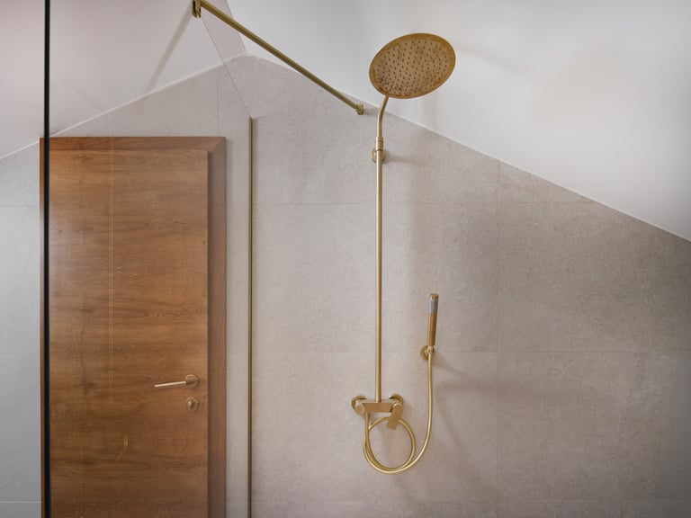 Modern brass shower system mounted on white wall with rainfall showerhead and handheld spray, next to wooden door
