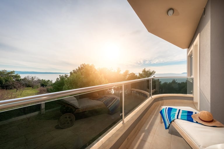A modern balcony overlooking a lake at sunset, with a striped beach towel and hat draped over a lounge chair
