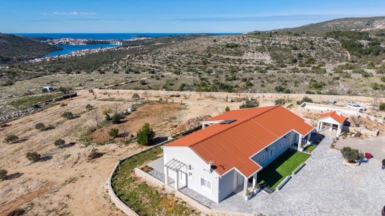 Aerial view of a modern white house with terracotta roof on rocky hillside overlooking a Mediterranean bay with blue water