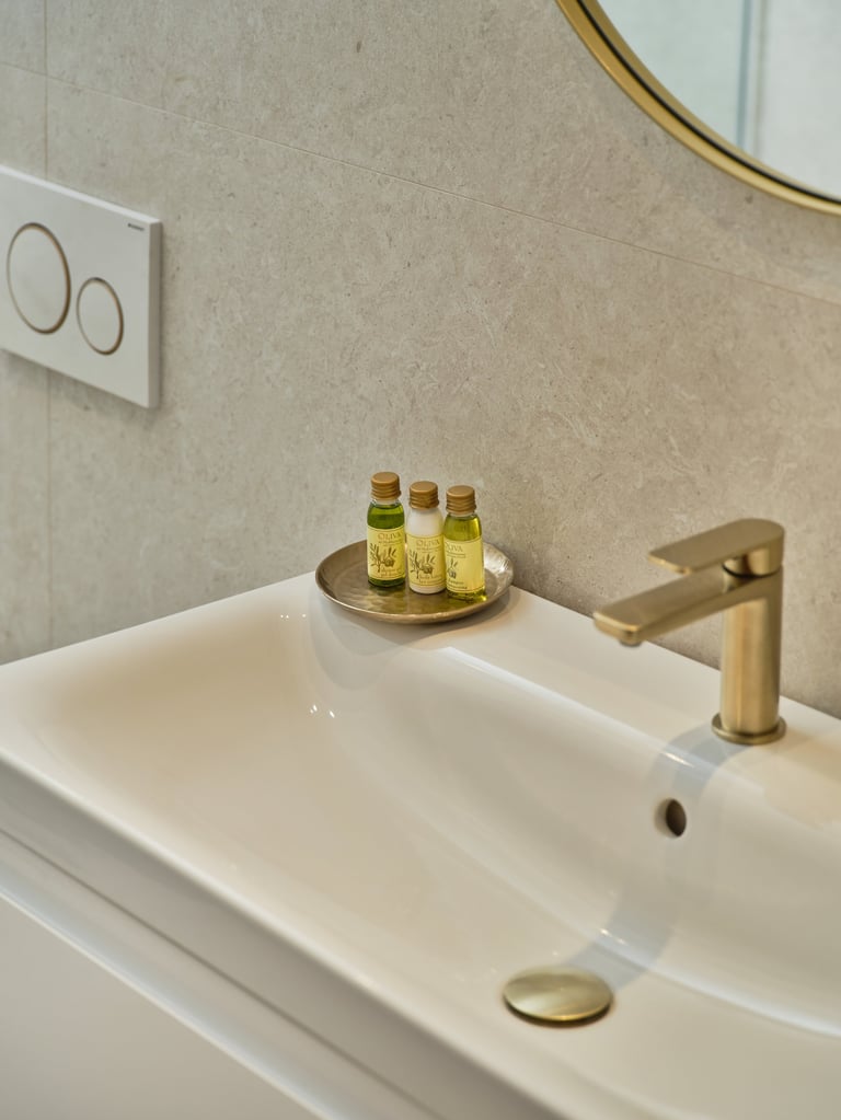 Modern bathroom sink with brass faucet, decorative bottles on tray, and round gold mirror on beige tile wall