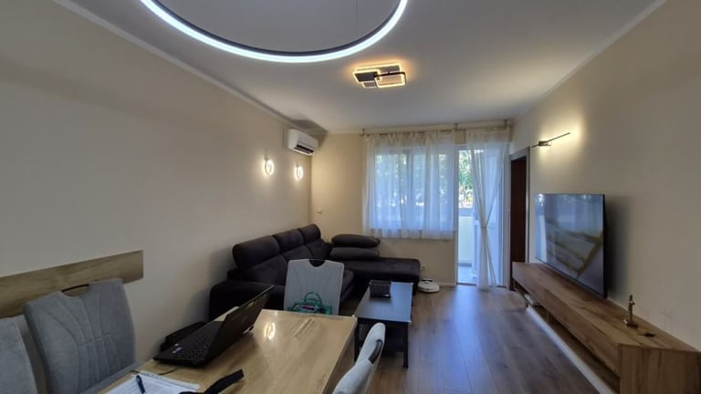 Modern living room with black sectional sofa, wooden coffee table, wall-mounted TV, glass balcony doors, and contemporary ceiling lighting