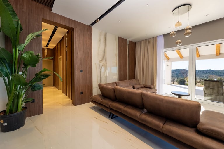 Modern living room with brown leather sofa, wood paneling, pendant lights, large windows overlooking mountains, and green plant accent