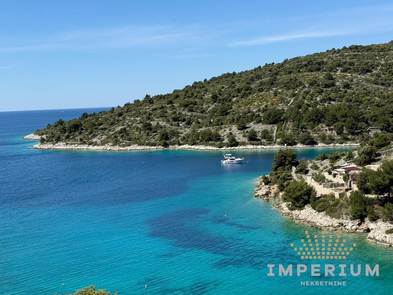Turquoise Mediterranean cove with forested hillside, white boat, and rocky coastline under clear blue sky