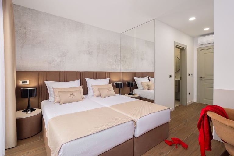Modern bedroom with two twin beds, white bedding, wooden headboard, bedside tables with lamps, full-length mirror, and wooden flooring