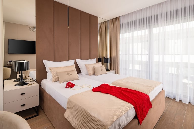 Modern bedroom with beige and brown tones, featuring a bed with red throw, wall-mounted TV, beige nightstand, and large windows with sheer curtains