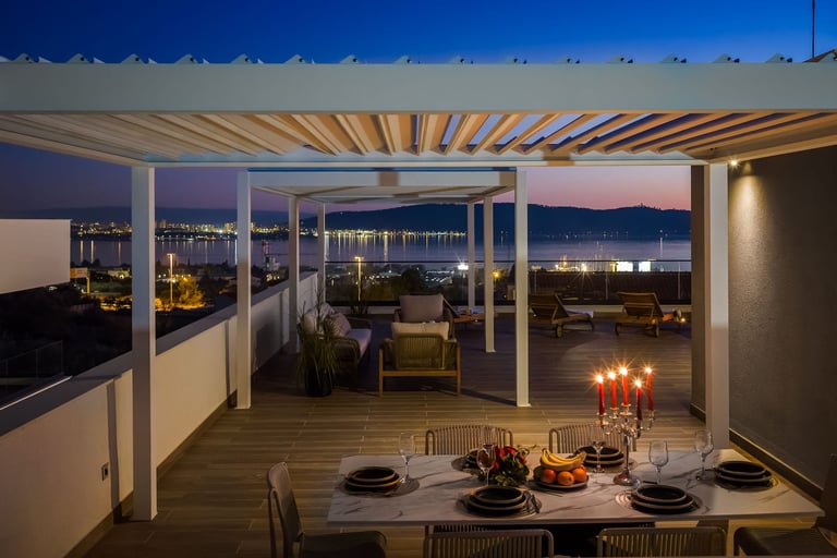 Modern rooftop terrace with dining table at dusk, featuring lounge seating, pergola, and illuminated city views at night