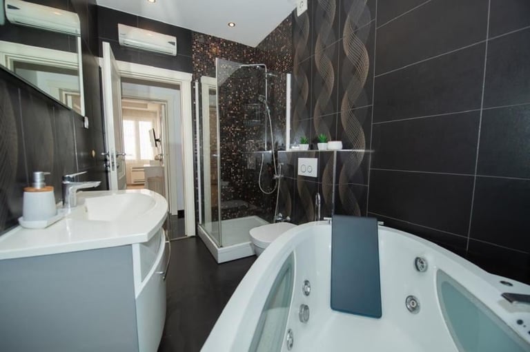 Modern bathroom with dark tile walls, white basin sink, glass shower enclosure, and jacuzzi bathtub with blue accent panel