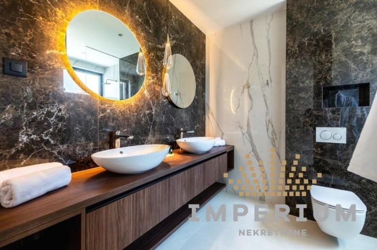 Modern luxury bathroom with dark marble walls, round illuminated mirror, white vessel sink, wood vanity, and decorative tiles