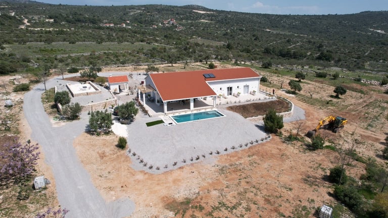 Aerial view of a modern white and red-roofed villa with swimming pool surrounded by rocky terrain and Mediterranean hills