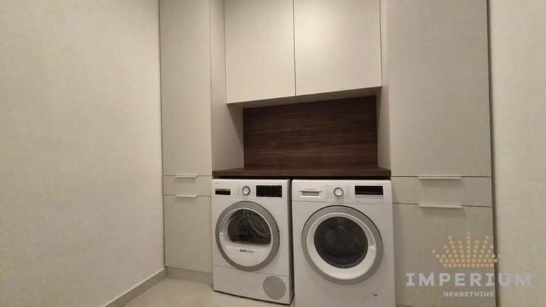 Two white front-load washing machines positioned side by side under a dark wooden shelf in a white laundry room