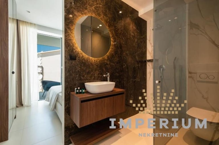 Modern bathroom with dark marble walls, round illuminated mirror, wooden vanity with white vessel sink, glass shower enclosure, and view to bedroom