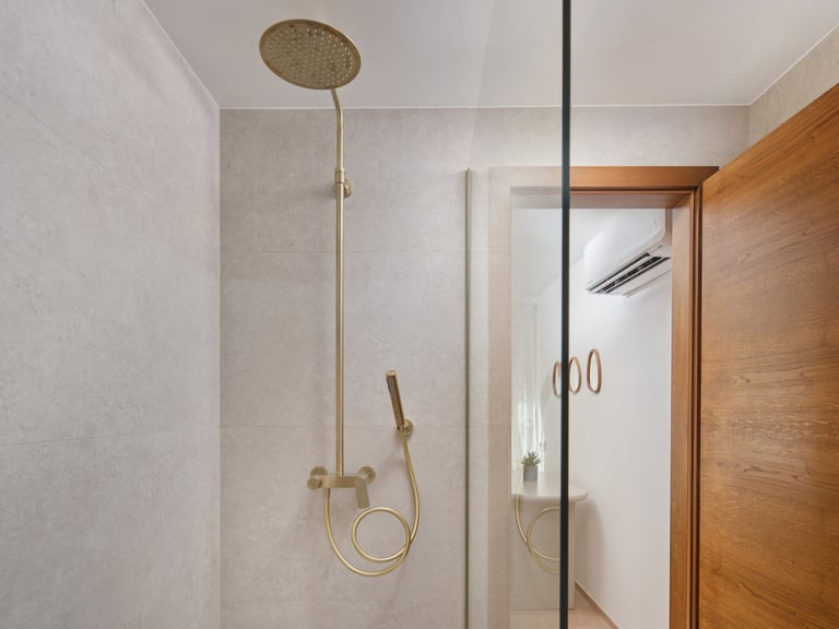 Modern bathroom shower with brass rainfall showerhead and hand sprayer mounted on gray tile walls with glass partition and wooden door visible