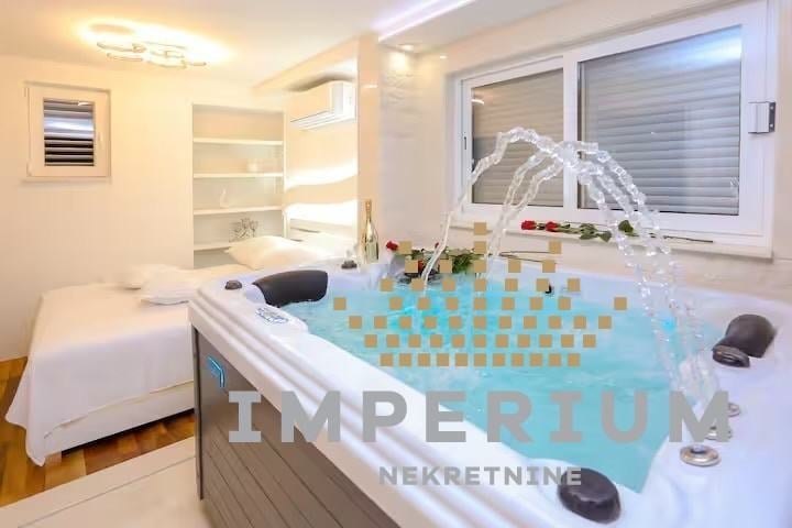 Modern bathroom with illuminated bathtub featuring blue mosaic tiles, massage jets, potted orchid plant, white cabinetry, and bed visible through doorway