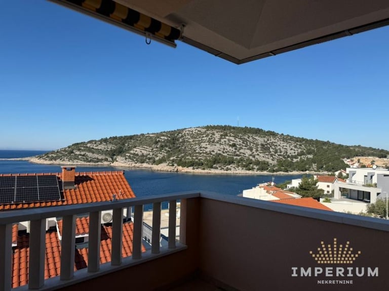 Elevated view from a balcony overlooking a Mediterranean coastal village with blue waters, terracotta roofs, and a forested hillside, with Imperium logo visible