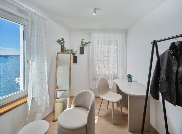 Bright minimalist room with white furniture, potted plants on wall, mirror, and sea view through window