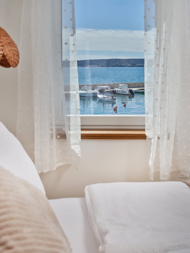 View from a bright bedroom with white sheer curtains framing a window overlooking boats and calm blue Mediterranean sea