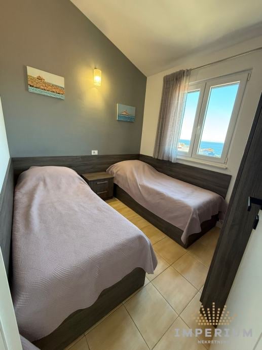 Small bedroom with two twin beds with purple bedding, grey walls, sea view window, pendant light, and beige flooring.