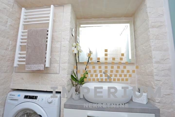 Modern bathroom with white washing machine, wall-mounted towel rack, white sink, and window with geometric tile pattern