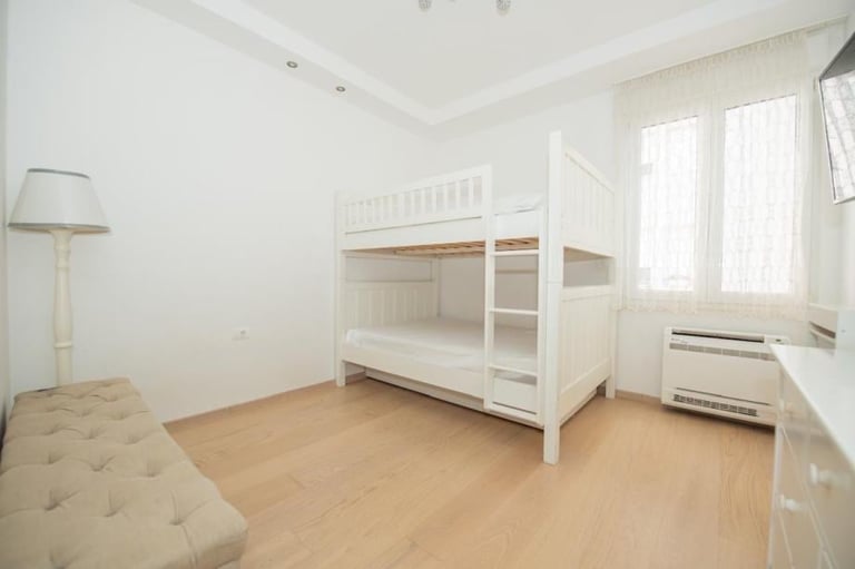 Bright bedroom with white bunk bed, wooden floor, bench, floor lamp, and windows with natural light