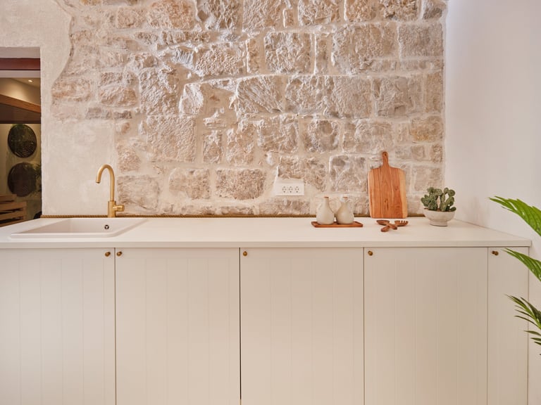 Modern kitchen counter with white cabinetry, sink, and brass faucet, backed by a rustic stone tile wall with decorative items
