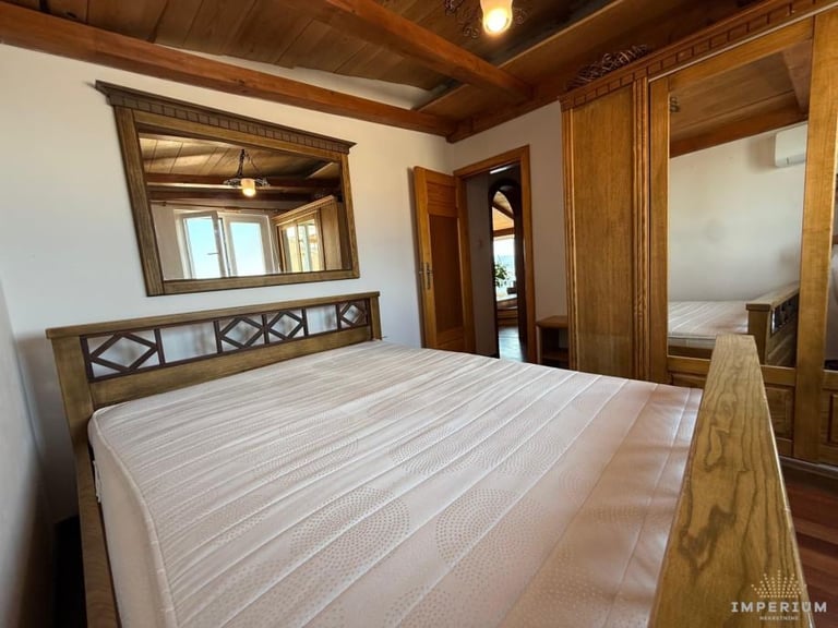 Spacious bedroom with wooden bed frame, white striped mattress, wooden ceiling beams, and a large mirror above the headboard