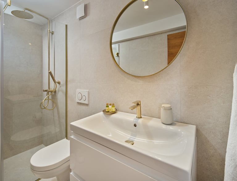 Modern bathroom with white vanity, round gold mirror, rainfall showerhead, and neutral tile walls