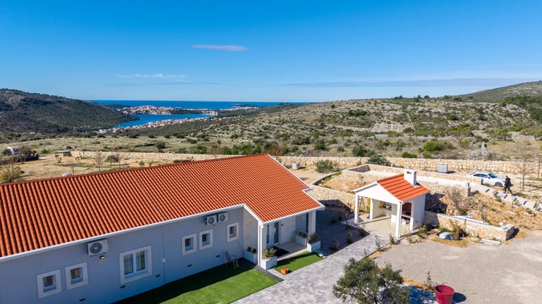 Aerial view of a white Mediterranean-style house with red terracotta roof in rocky coastal landscape overlooking blue sea