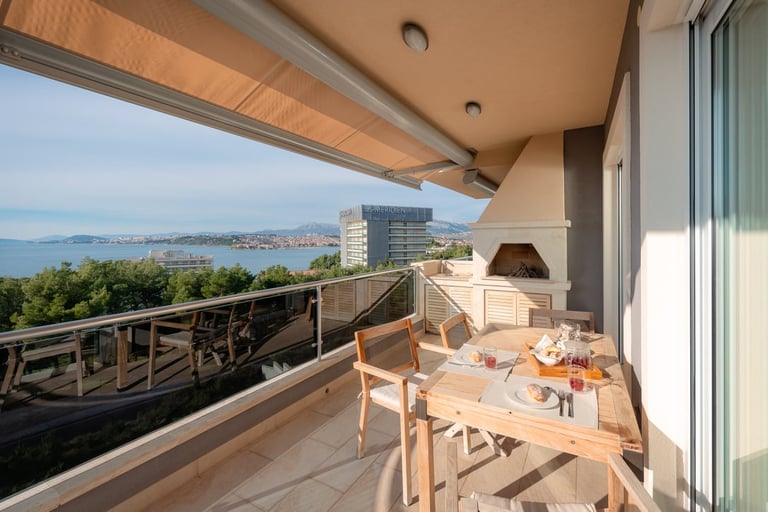 Modern apartment balcony with wooden dining furniture overlooking a coastal bay with islands and mountains in the distance