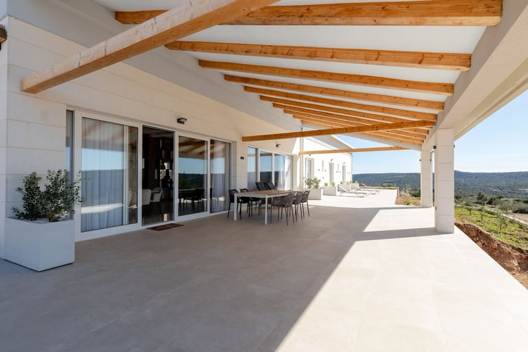 Modern house with expansive covered patio, wooden beam ceiling, large glass doors, outdoor dining table, and panoramic countryside views