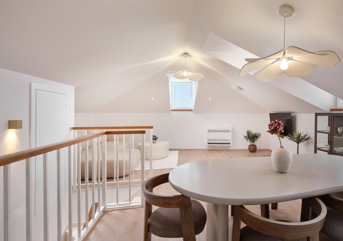 Modern loft space with white round dining table, wooden chairs, staircase railing, and ceiling fan under sloped roof
