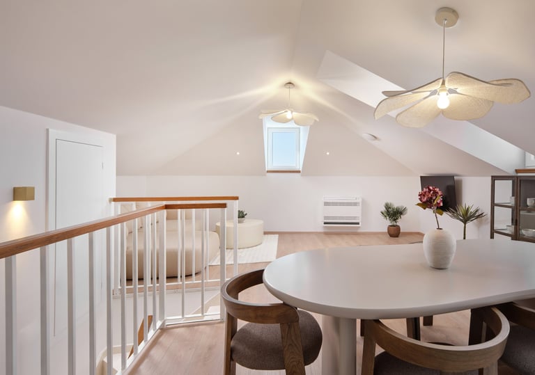 Modern loft space with white round dining table, wooden chairs, staircase railing, and ceiling fan under sloped roof