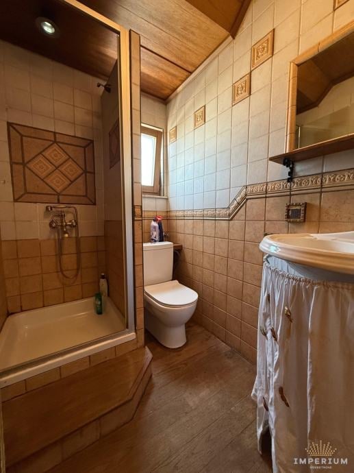 Warm-toned bathroom with beige tile walls, wooden shower enclosure, toilet, sink with wooden base, and wooden ceiling.