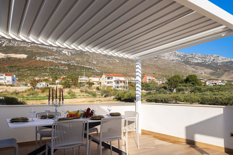Modern terrace with retractable white pergola, dining table, and chairs overlooking hillside village with mountains in background