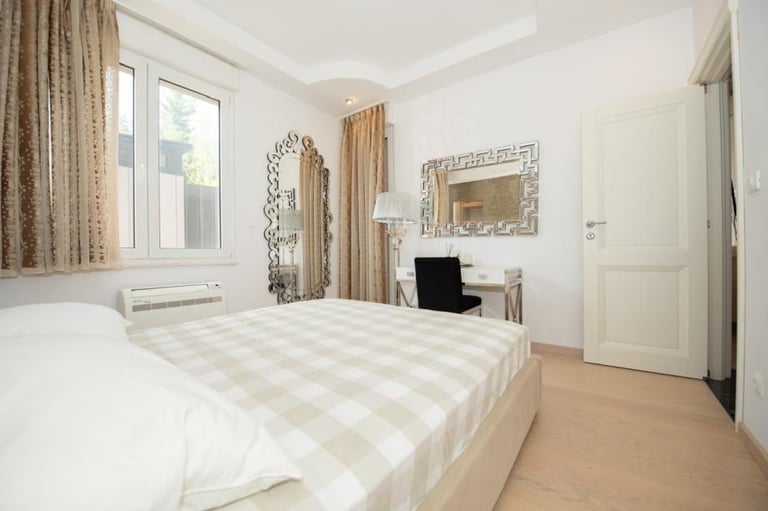 Bright modern bedroom with white and beige plaid bedding, tall windows with curtains, desk with monitor, ornate mirror, and light wood flooring