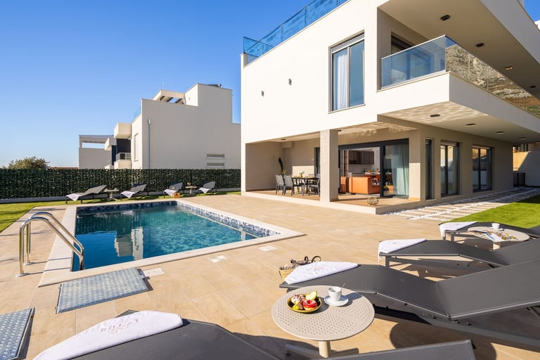 Modern white villa with swimming pool, loungers, and patio seating overlooking manicured garden