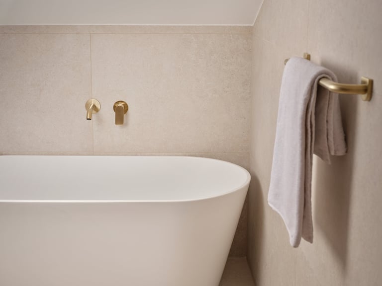 Modern beige bathroom with white freestanding tub, brass fixtures, and towel rack with folded towels
