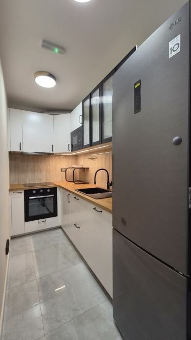 Modern galley kitchen with white and wood cabinetry, stainless steel refrigerator, black oven, and tiled flooring