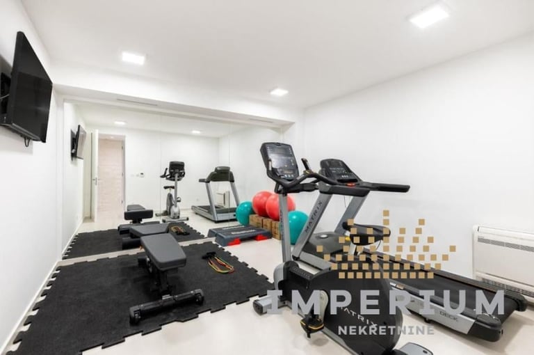 Modern home gym with exercise equipment including treadmills, stationary bikes, dumbbells, and black foam flooring in a bright white room