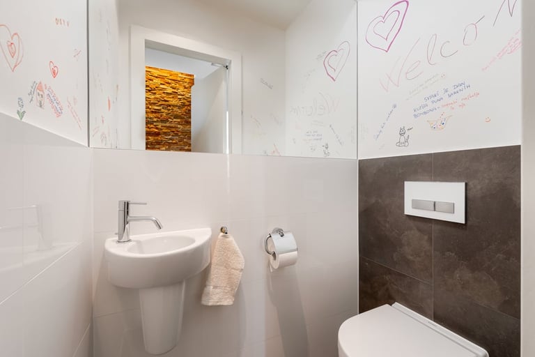 Modern bathroom with white walls featuring child-drawn doodles, round white sink with towel, chrome fixtures, dark tile accent, and toilet visible on right side