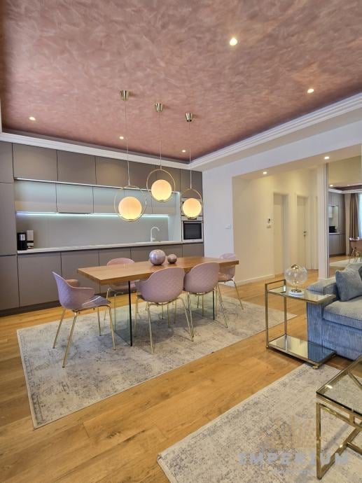 Modern apartment interior with open kitchen, dining area with wooden table and purple chairs, hardwood floors, and textured pink ceiling with pendant lights
