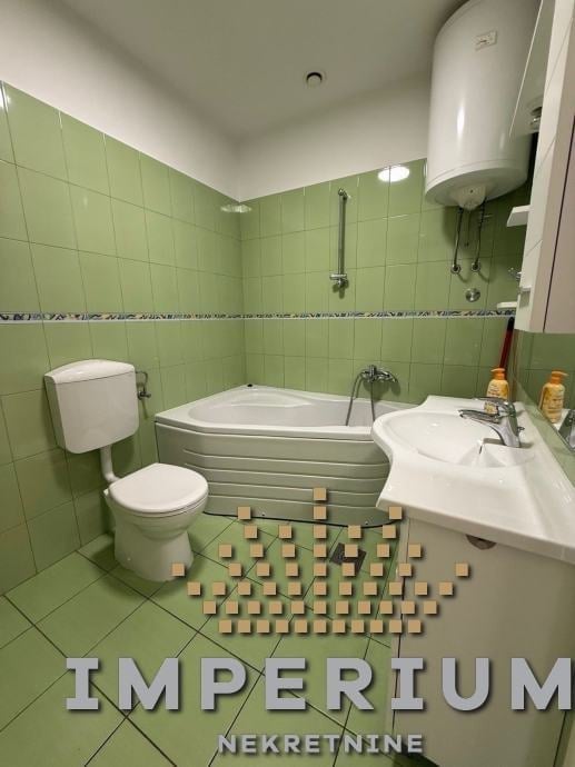 Bathroom with green tiles, white toilet, bathtub, sink, and wooden blocks on floor arranged in pattern with Imperium Nekretnine text overlay